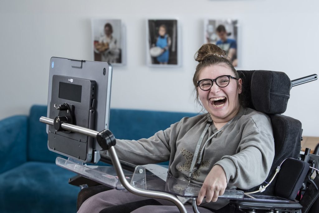 Eine junge Frau im Rollstuhl lacht, vor ihr ein Sprachcomputer mit Augensteuerung